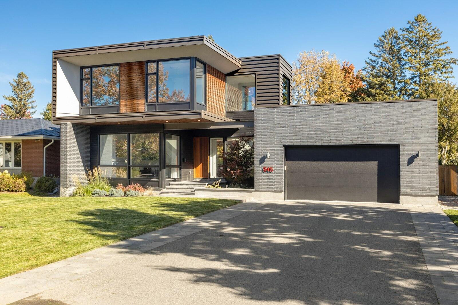 A modern two-story house with a brick facade, large windows, and a driveway, surrounded by fall foliage