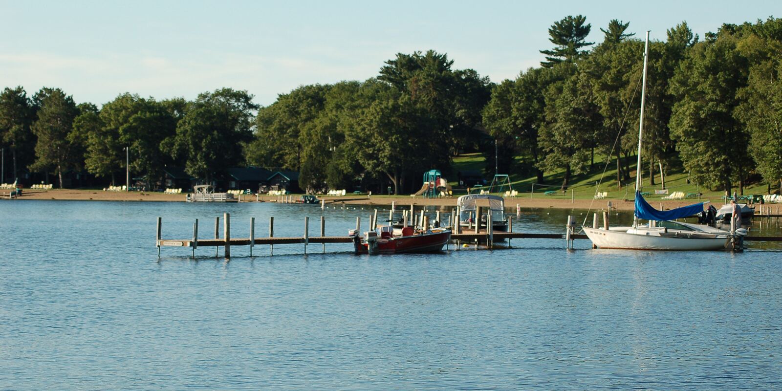 A dock with boats moored on a Minnesota lake, with a sandy beach, playground, and tree-lined shore in the background