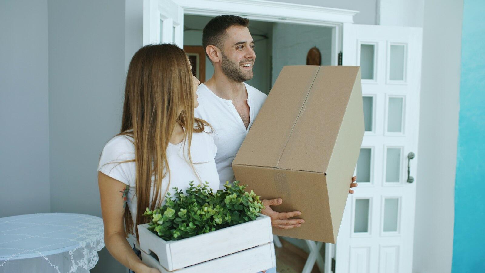 A couple carrying a moving box and a small plant into their new home